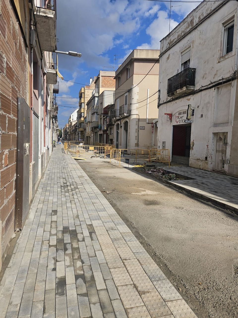 Calle Sant Pere en obras mirando hacia el final: acera nueva de adoquín gris con franja podotáctil, calzada con la base extendida y vallas amarillas