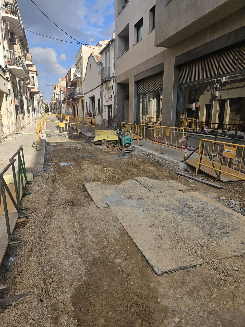 Tramo de la calle Sant Pere todavía levantado, con planchas metálicas sobre el terreno, una compactadora y vallas junto a la acera