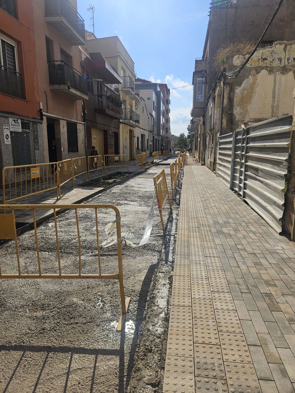 Calle Sant Pere con vallas amarillas, una acera nueva con franja podotáctil y la calzada todavía sin pavimento definitivo